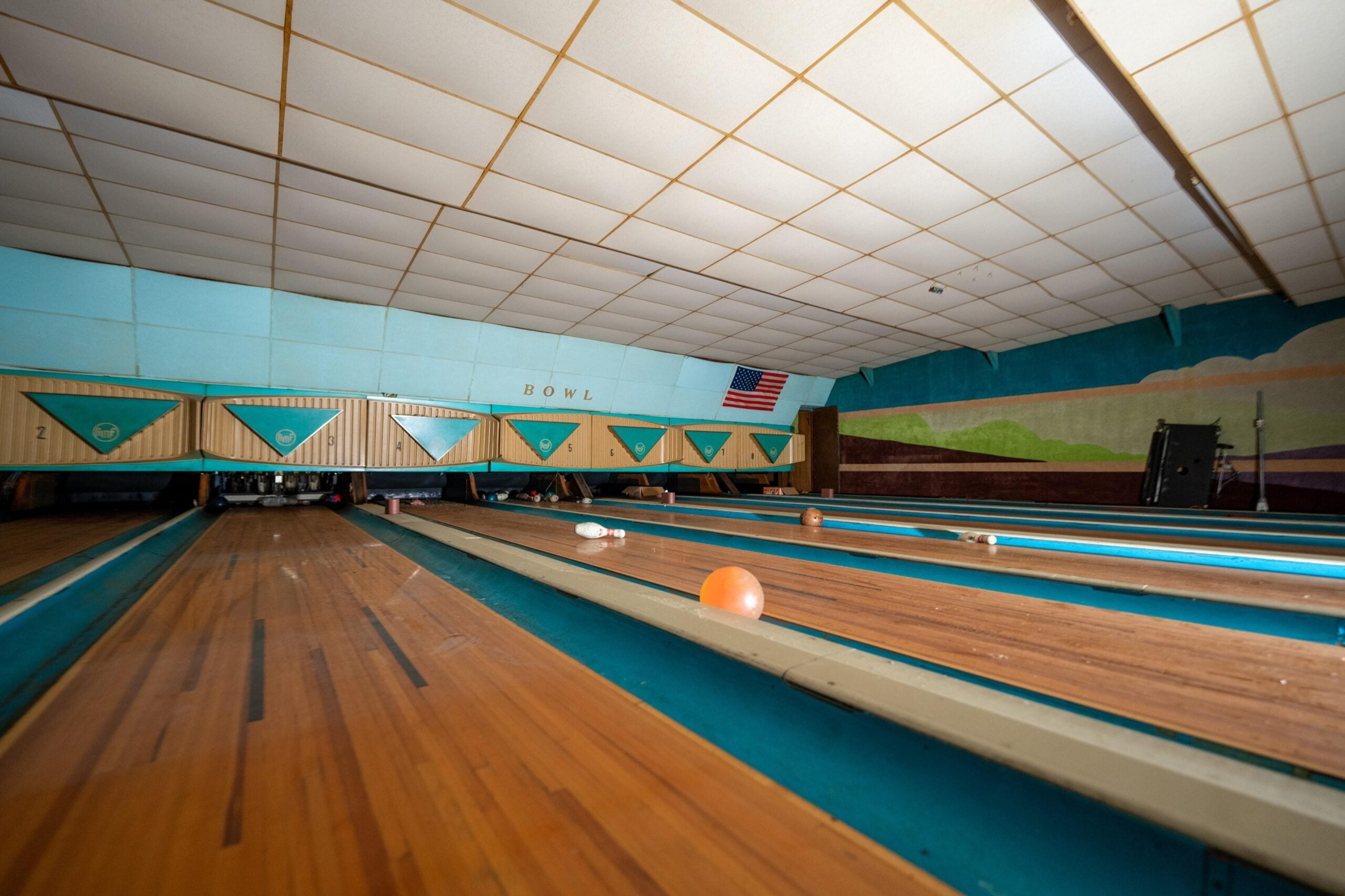 An empty vintage bowling alley with wooden lanes, a few scattered bowling balls, turquoise accents, and a mural with an American flag on the back wall. The ceiling has a grid pattern of tiles.