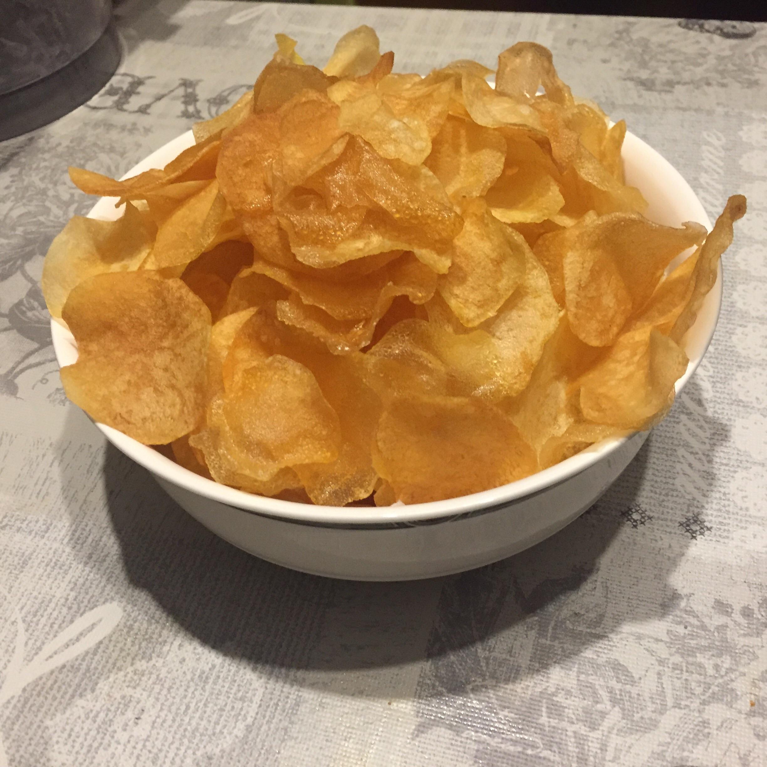 A white bowl filled with golden, crispy potato chips sits on a patterned tablecloth.