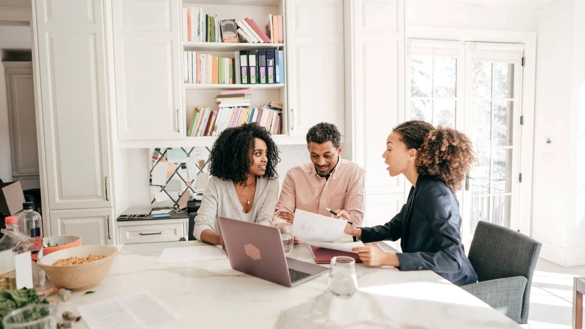 Three people sit at a kitchen table with a laptop and papers, having a discussion. Bookshelves and bright windows are in the background, and the atmosphere appears friendly and collaborative.