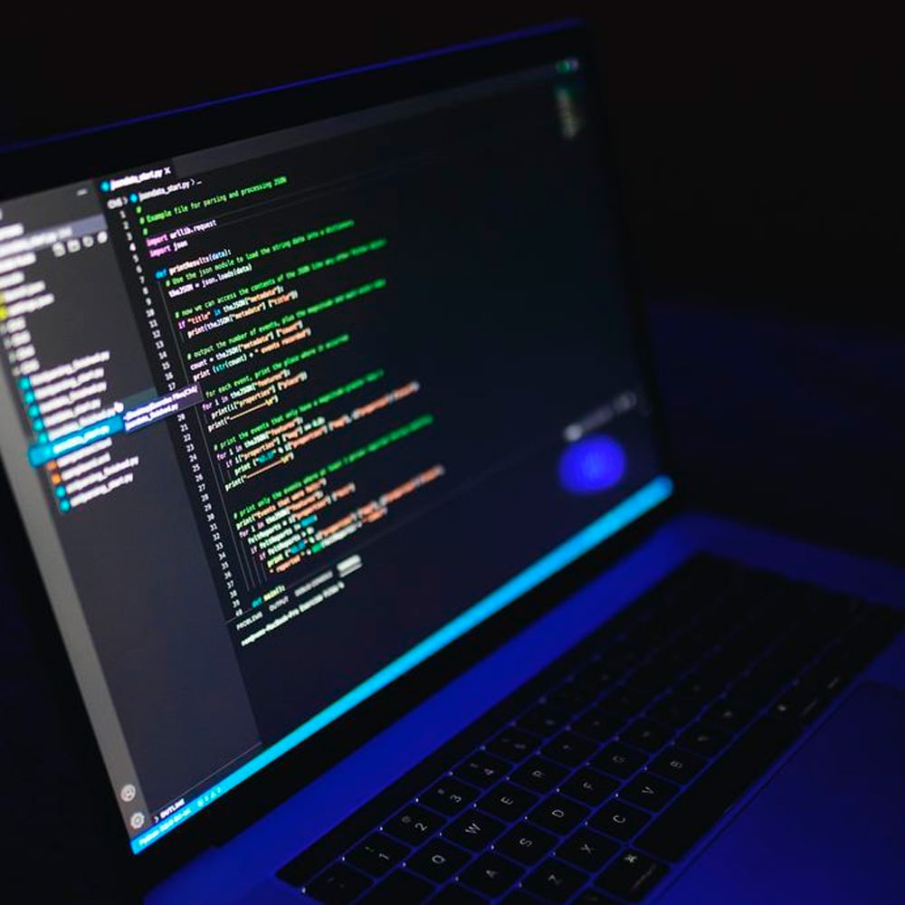 A laptop displaying lines of colorful code on a dark-themed text editor screen, illuminated by a blue glow in a dimly lit environment.