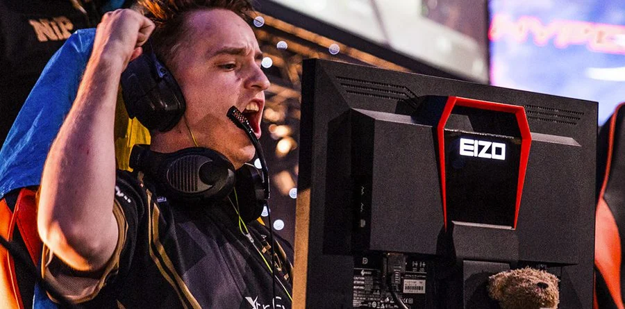 A person wearing headphones and a gaming jersey raises a fist and shouts in excitement while sitting in front of a computer monitor at an esports event.
