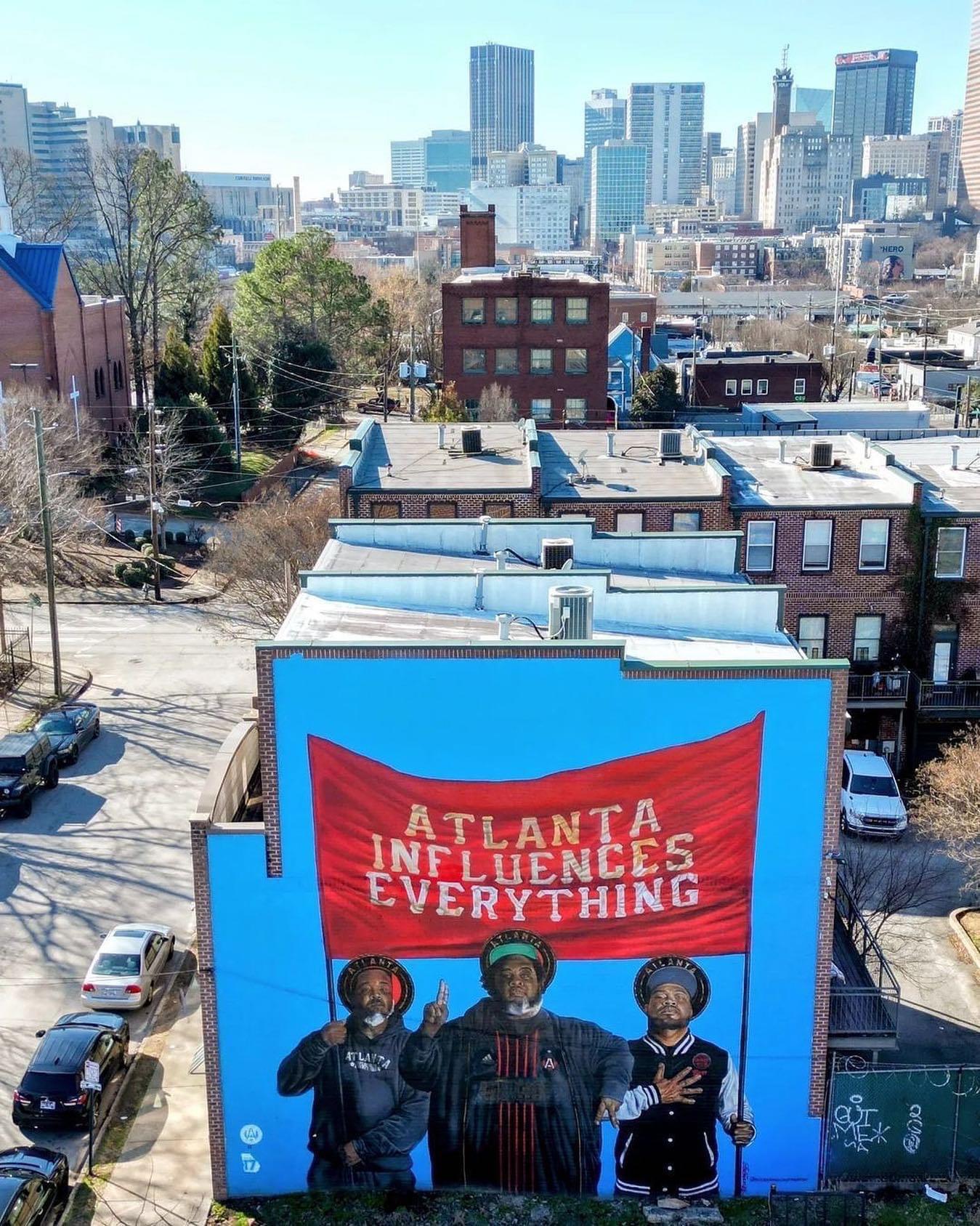 A mural on a building in Atlanta shows three people holding a large red banner that reads "Atlanta Influences Everything," with the city skyline visible in the background.