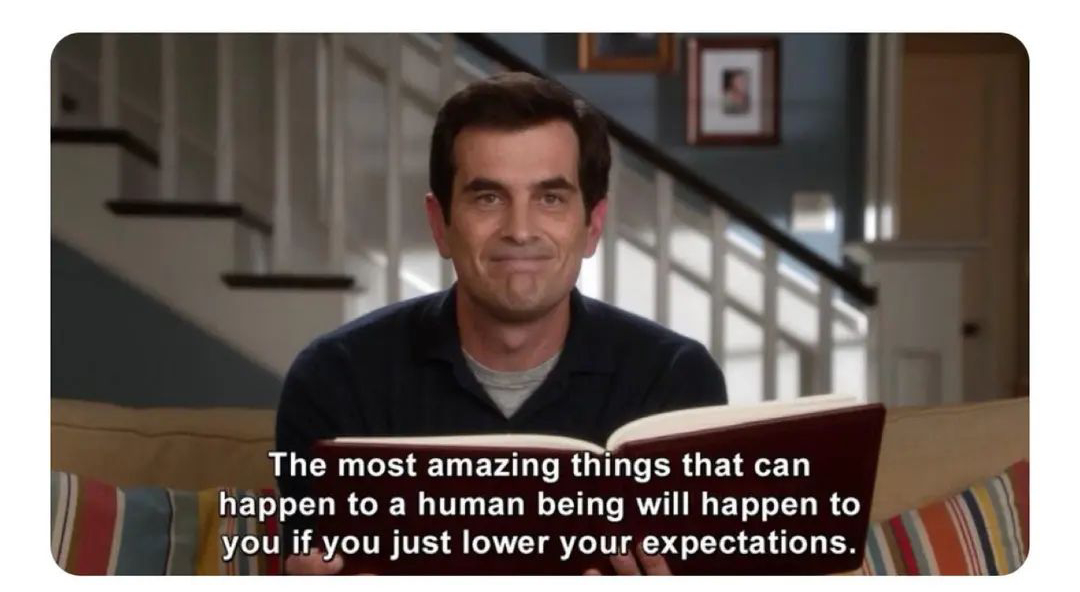 A man sits on a couch holding a book, smiling, with stairs in the background. The caption reads: "The most amazing things that can happen to a human being will happen to you if you just lower your expectations.