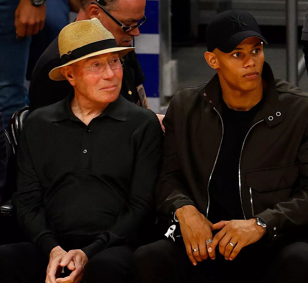 Two men sit side by side at an indoor event. The older man on the left wears glasses, a straw hat, and a black shirt. The younger man on the right wears a black cap, a dark jacket, and looks to the side.