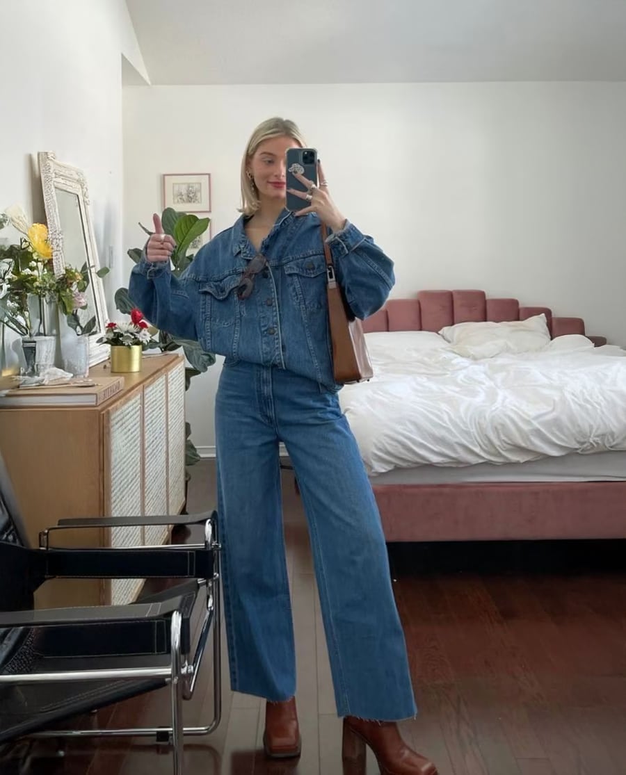 A woman stands in a bedroom taking a mirror selfie, wearing a denim jacket and jeans with brown boots. She holds up a peace sign, and a bed, flowers, and a chair are visible in the background.