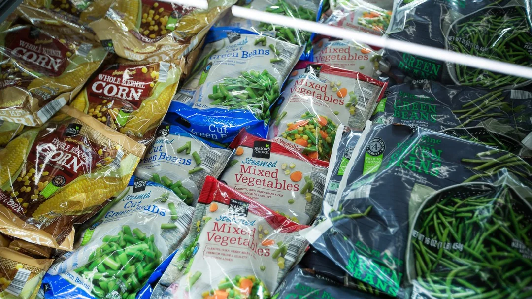 Bags of frozen vegetables, including corn, green beans, and mixed vegetables, are arranged on shelves in a grocery store freezer section.