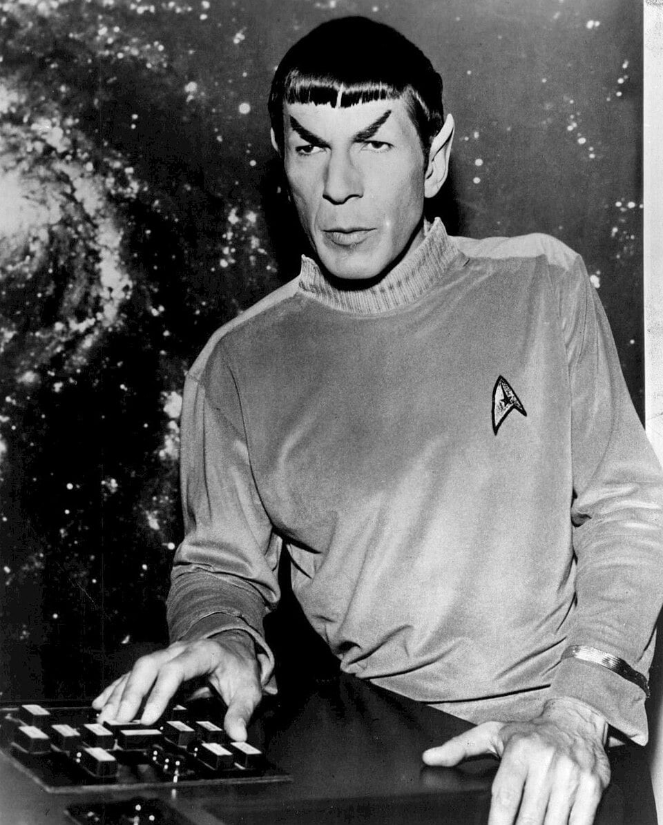 A person dressed as a science officer from Star Trek, wearing a long-sleeved shirt with an insignia, pointed ears, and straight-cut bangs, stands at a control panel with a galaxy in the background.