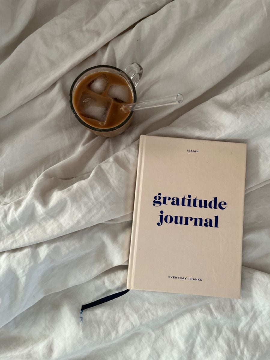 A beige gratitude journal and a glass of iced coffee with a straw rest on a white, rumpled bedsheet.
