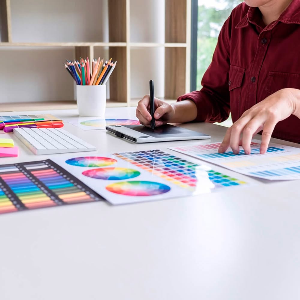A person in a red shirt uses a digital drawing tablet at a desk covered with color swatches, charts, a keyboard, sticky notes, and a cup filled with colored pencils, suggesting graphic design work.
