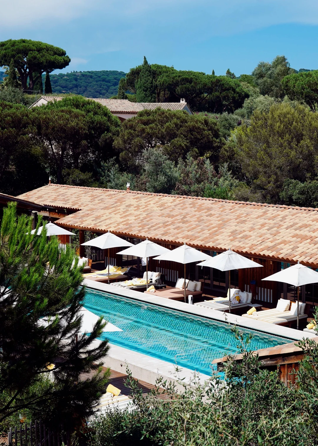 A rectangular outdoor pool with white umbrellas and lounge chairs sits beside a terracotta-roofed building, surrounded by lush green trees and distant hills under a blue sky.