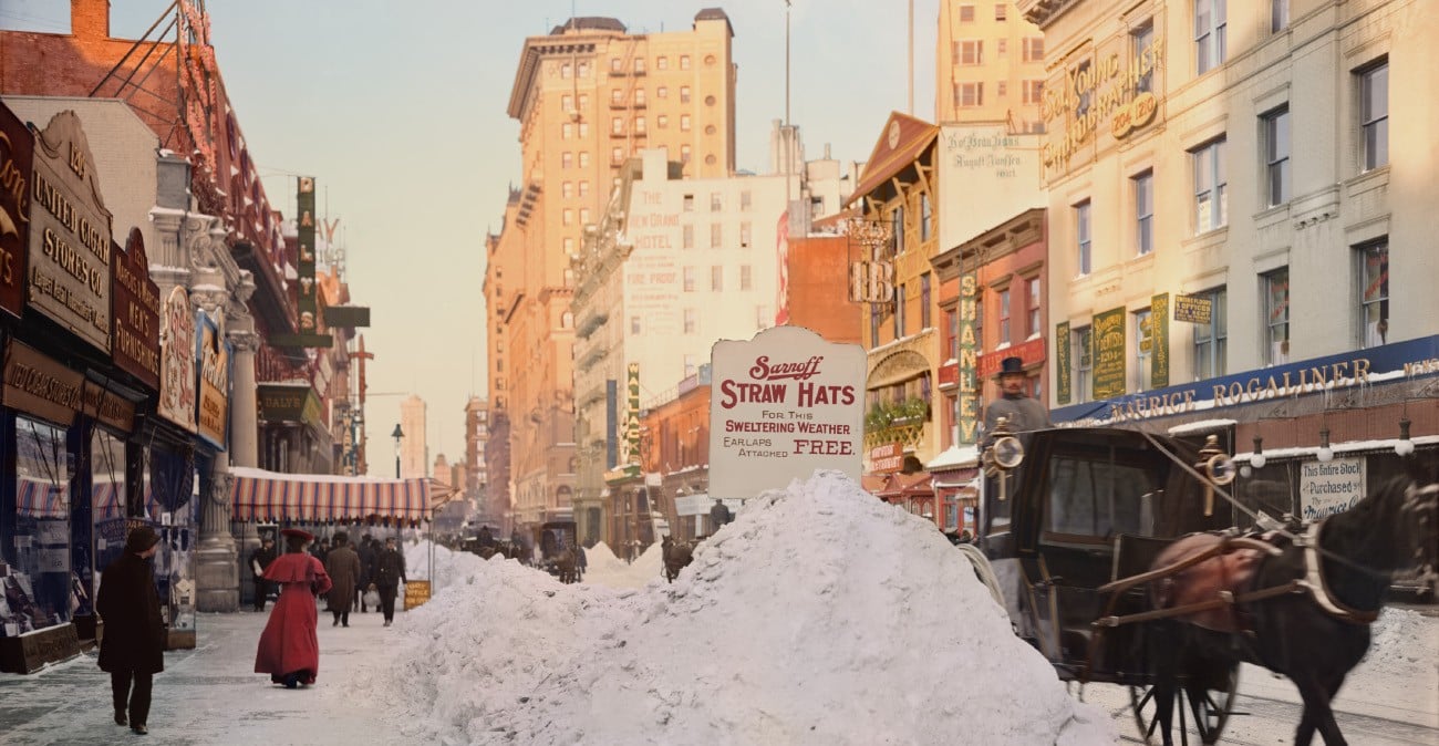 A busy city street in winter, lined with shops and tall buildings. Pedestrians walk past a large snow pile while a horse-drawn carriage travels by. Shop signs and advertisements are visible along the storefronts.