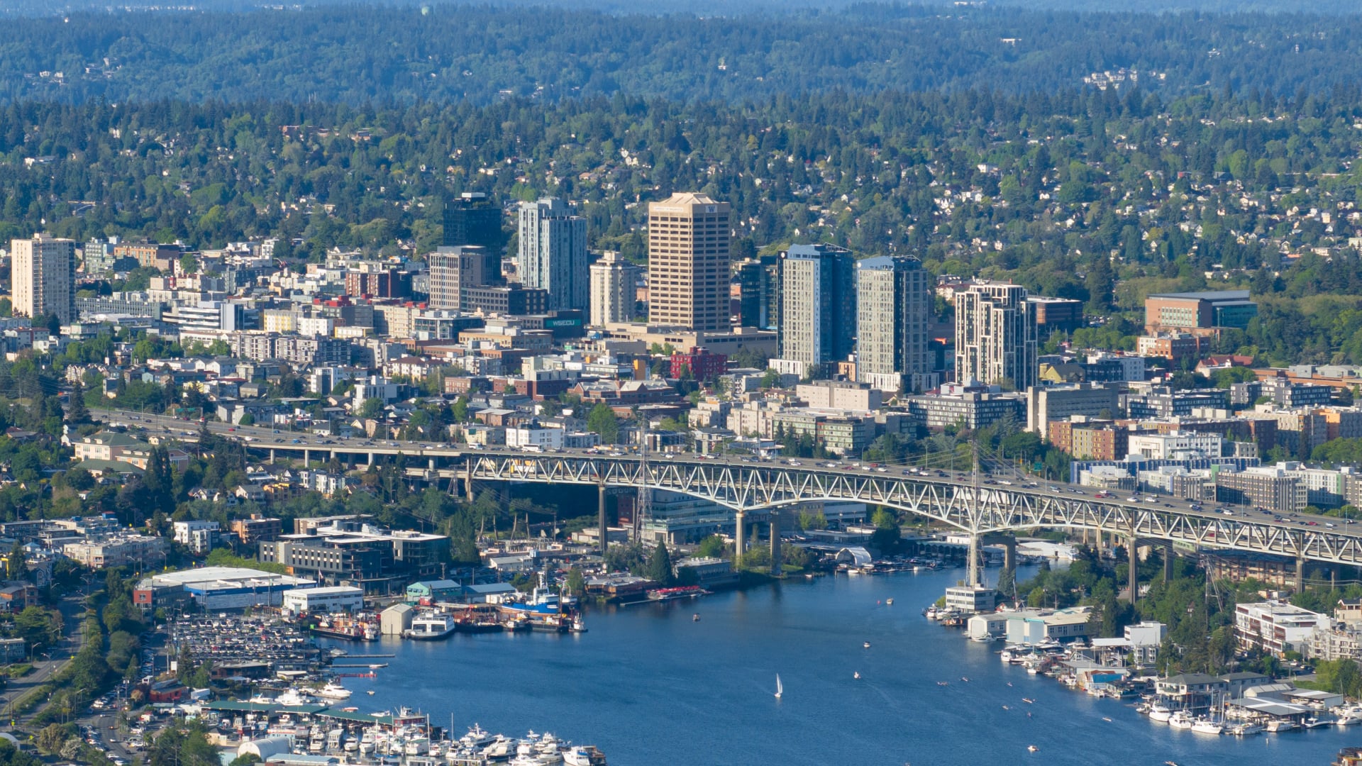 Aerial view of a city with tall buildings, a large bridge spanning a river or lake, and boats docked in the water below; the background includes a forested area and distant hills.