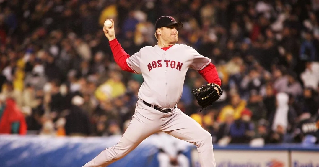 A baseball player in a Boston Red Sox uniform winds up to pitch during a game, with a blurred crowd in the background.