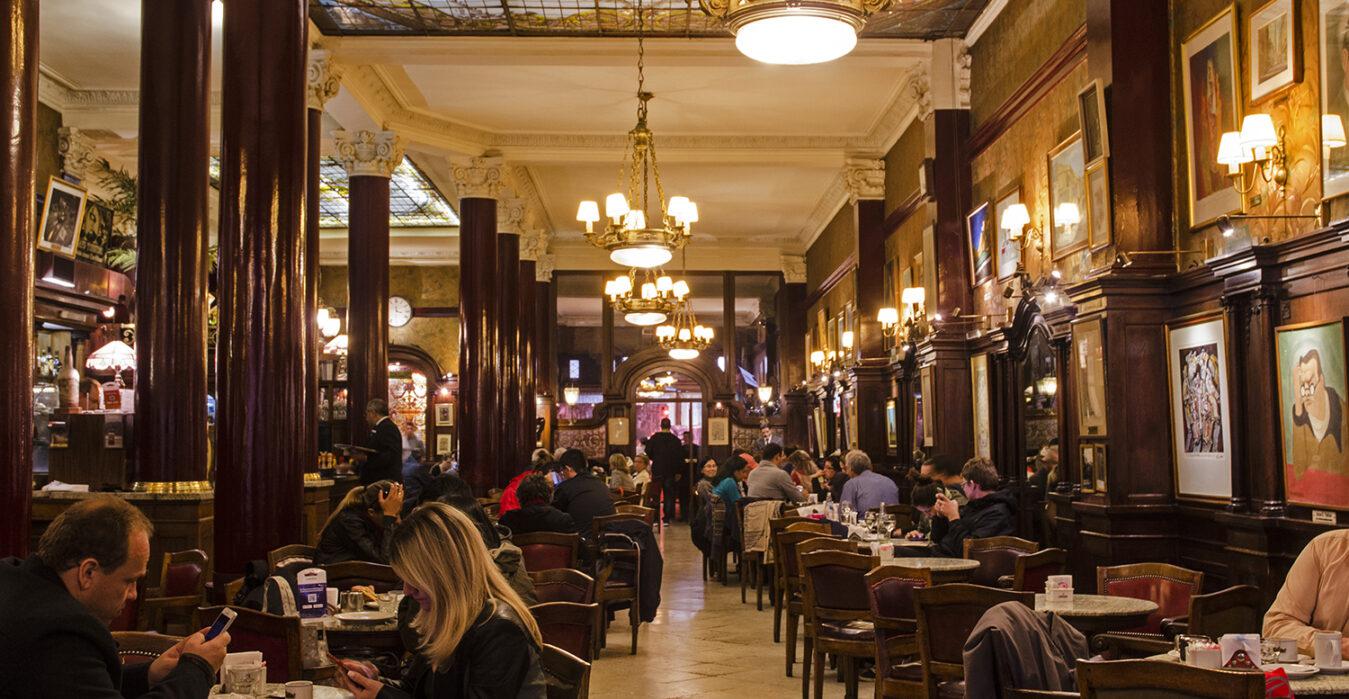 A cozy, elegant café with patrons seated at wooden tables, chandeliers hanging from a high ceiling, dark wood pillars, and artwork displayed on the walls. Warm lighting fills the lively, welcoming atmosphere.
