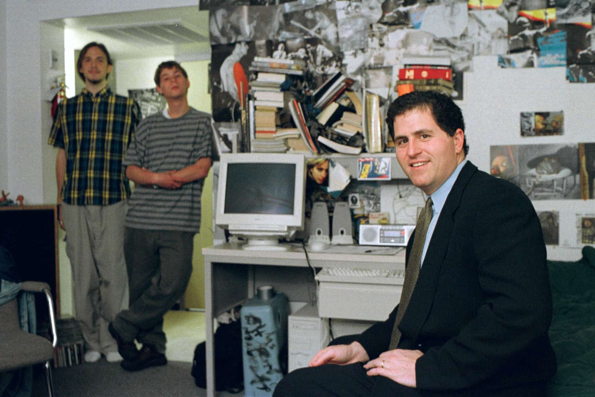 A man in a suit sits in front of a desk with a computer, while two casually dressed young men stand in the background. The room has posters and books stacked on a shelf above the desk.