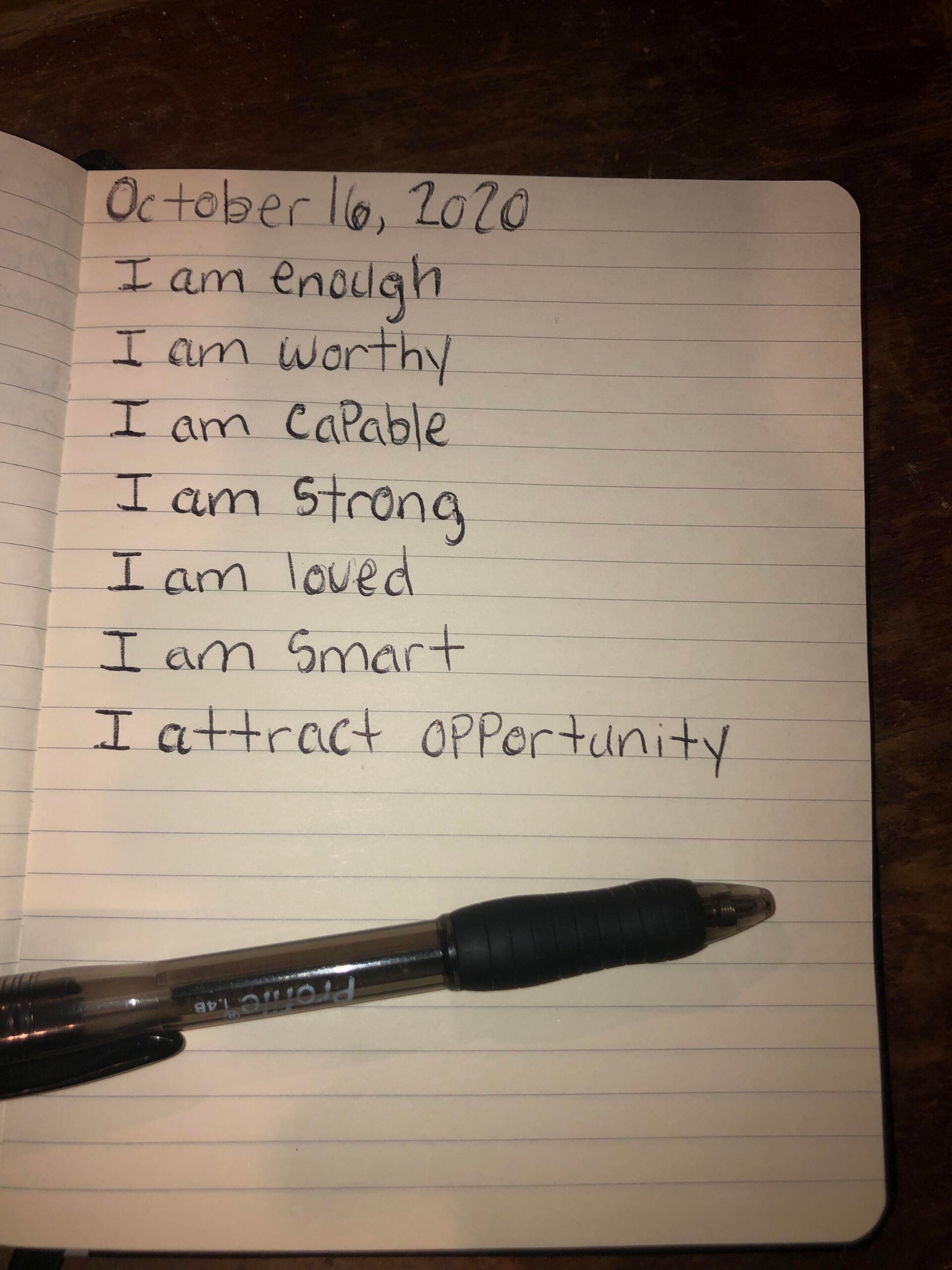 A notebook open to a page dated October 16, 2020, displaying handwritten affirmations such as "I am enough," "I am worthy," and "I attract opportunity." A black pen rests on the notebook.
