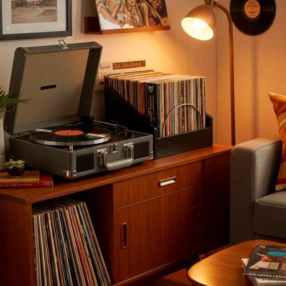 A cozy living room corner with a record player on a wooden cabinet, vinyl records in black crates, a small lamp, framed pictures on the wall, and a book and mug on a side table next to a sofa with striped pillows.
