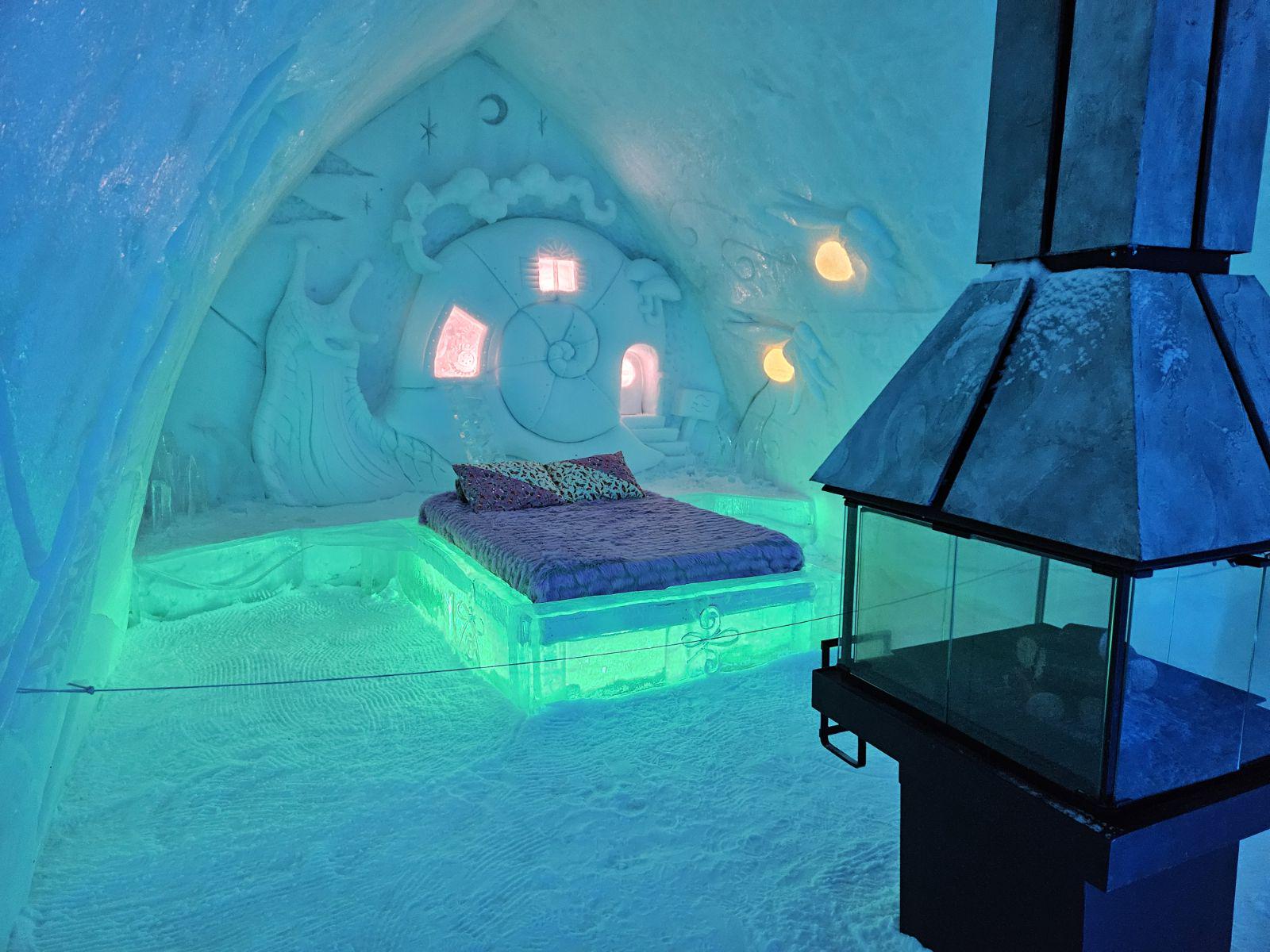 A cozy bed with glowing green lights sits in an ice hotel room, surrounded by icy walls carved with whimsical designs. A modern metal fireplace stands nearby under soft blue lighting.