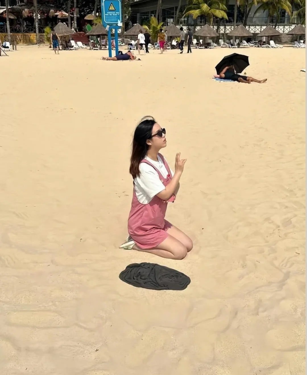 A woman in pink overalls and sunglasses kneels on sandy beach with hands in a prayer gesture. People relax in the background near umbrellas and palm trees. A dark shirt lies on the sand in front of her.