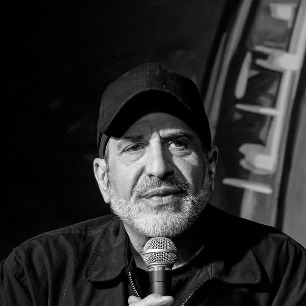 Black and white photo of an older man with a beard and a black cap, holding a microphone. He is wearing a dark jacket and appears to be speaking. The background is blurred with abstract shapes.