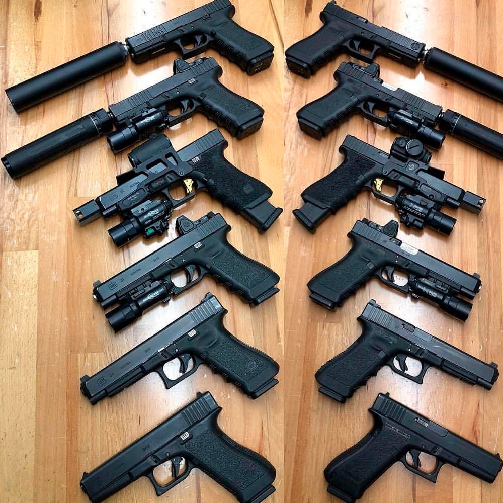 Twelve black handguns, some with attachments like suppressors, extended magazines, and sights, are arranged in two columns on a wooden floor.