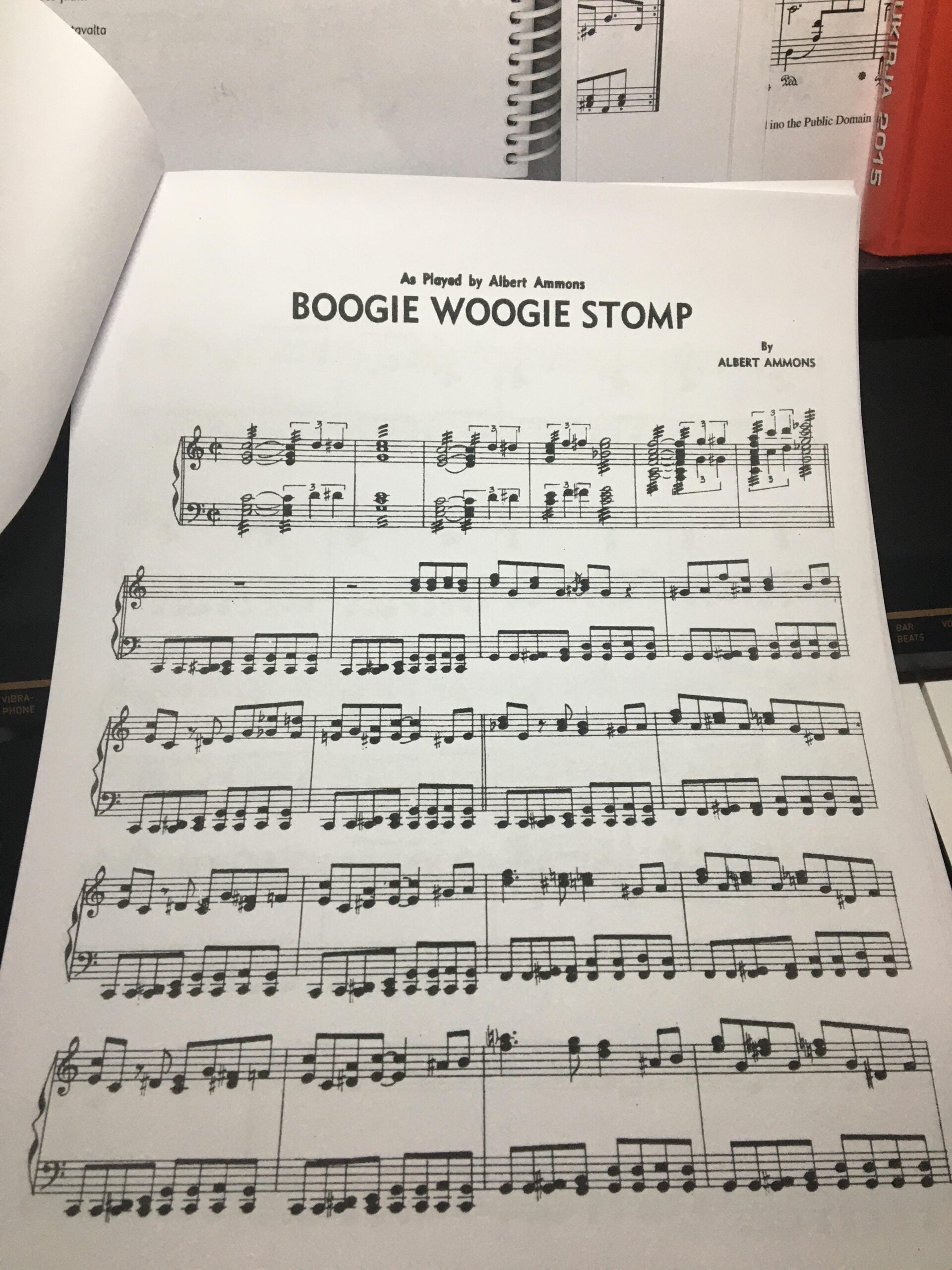 A sheet of piano music titled "Boogie Woogie Stomp" by Albert Ammons, with musical notation and two staves per system. The sheet rests on a piano with open books in the background.