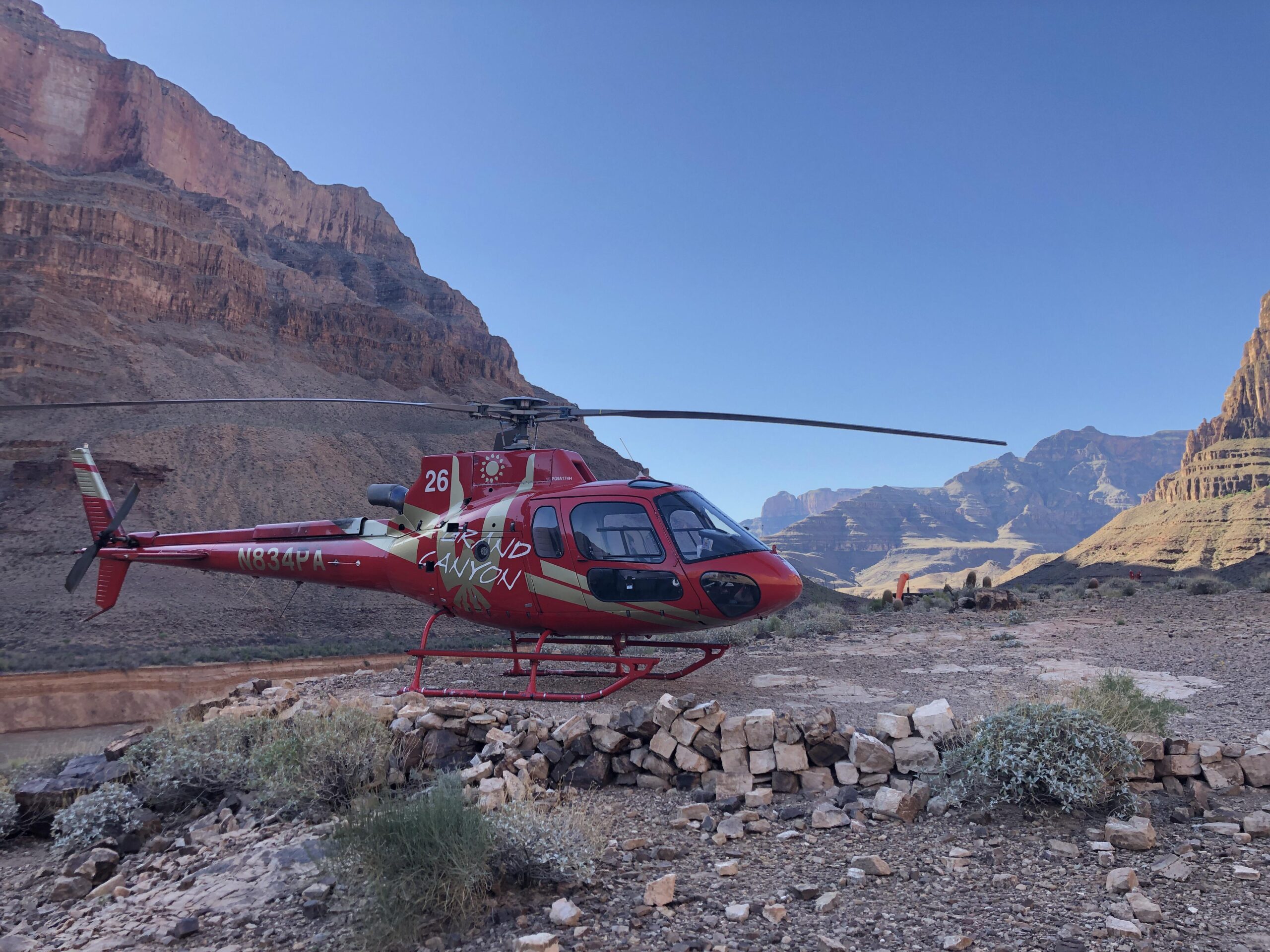 A red helicopter with "Grand Canyon" written on the side is parked on rocky terrain with stone piles, surrounded by steep canyon walls under a clear blue sky.