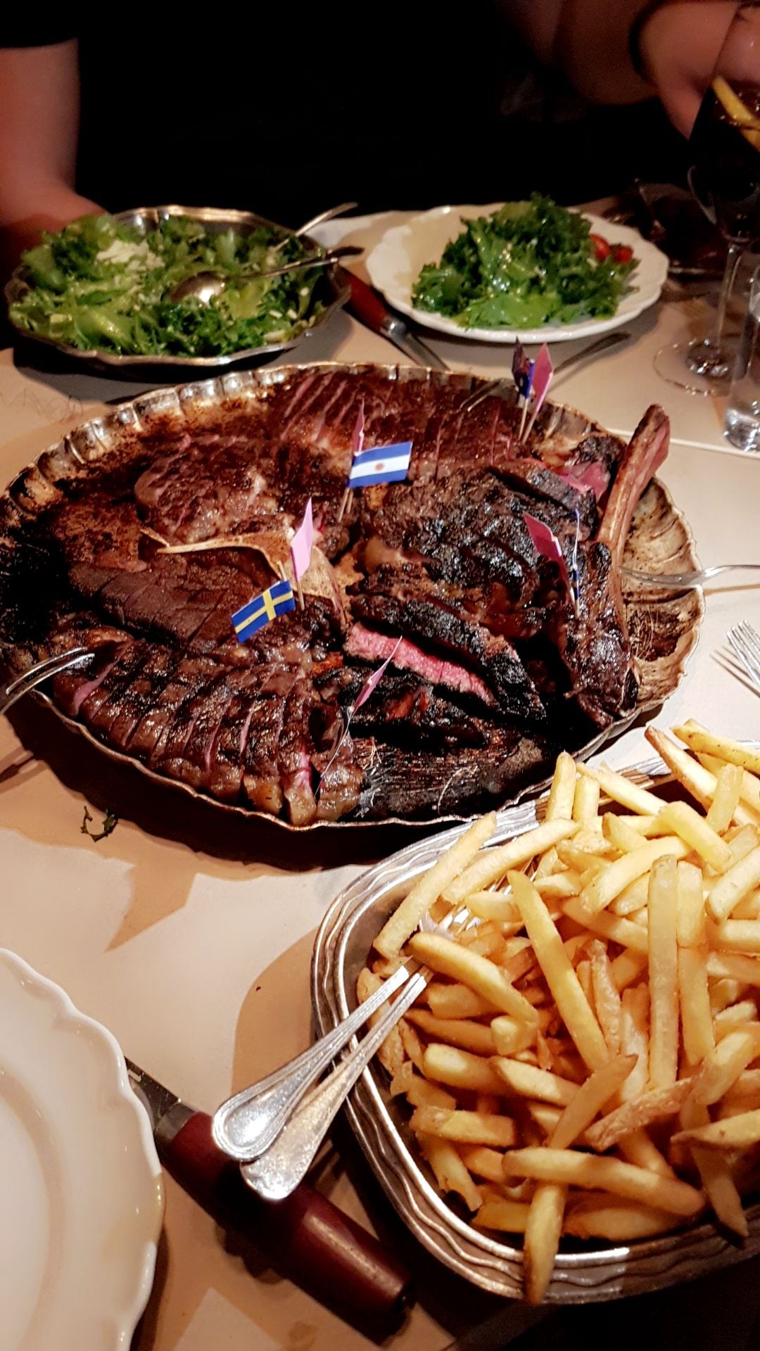 A platter of sliced steak with small flags, a dish of French fries, and plates of green salad on a restaurant table, with a person partially visible in the background.