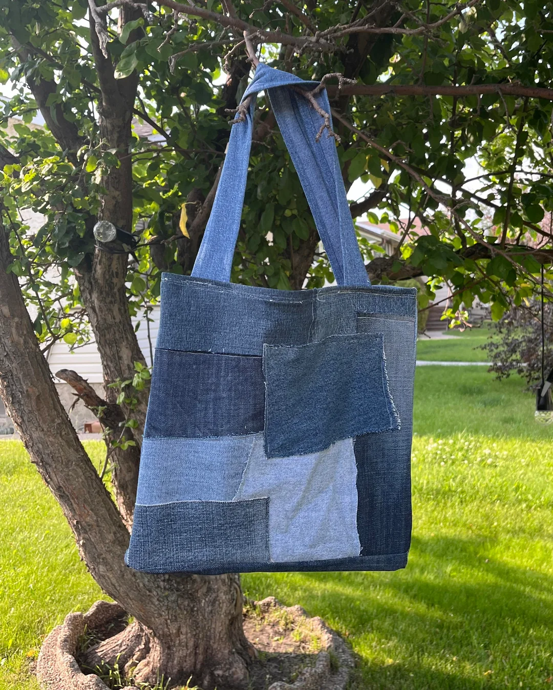 A handmade tote bag made from various shades of denim patches hangs from a tree branch in a sunny backyard with green grass and leafy trees.