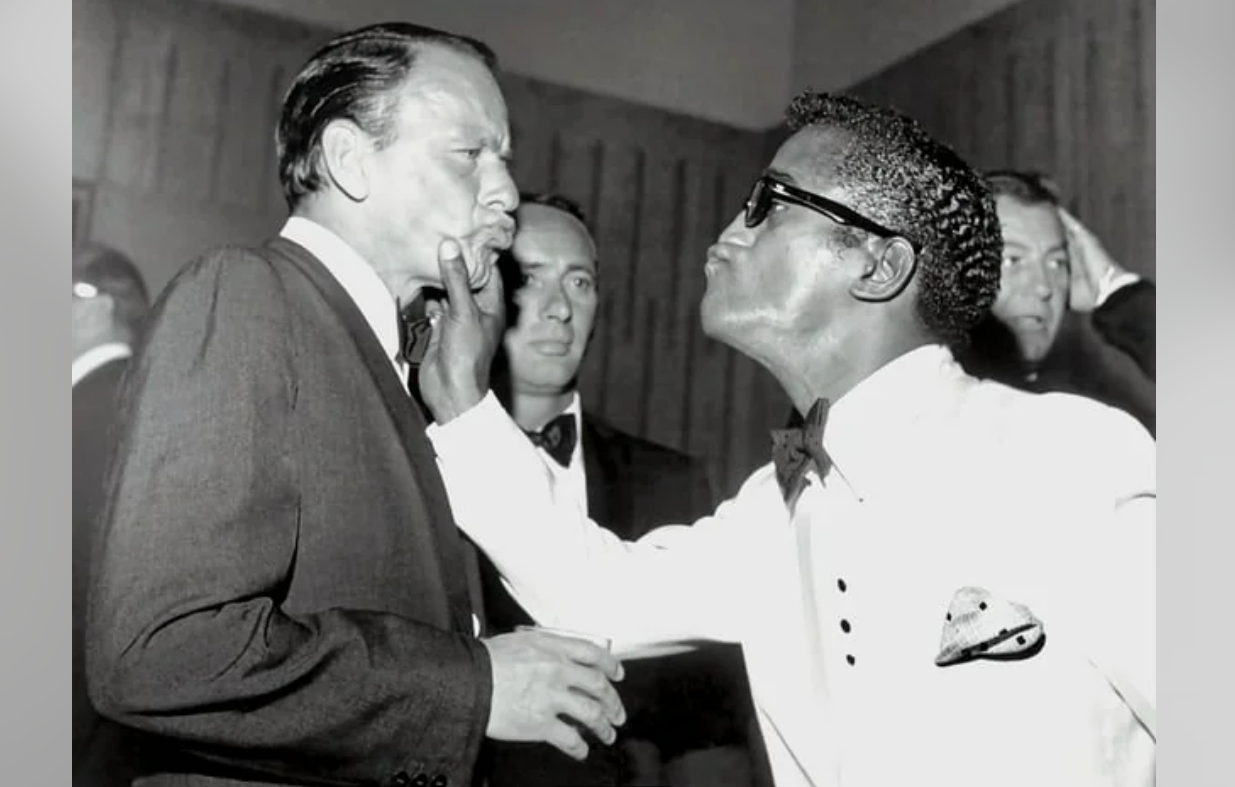 A Black man in a white suit and sunglasses playfully grabs a white man’s chin, leaning in as if to kiss him. Two other men in suits watch in the background. The scene appears light-hearted and humorous.