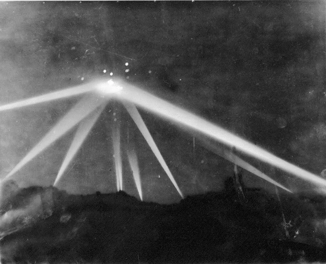 Black and white photo showing several beams of light converging in the night sky, likely from searchlights, with bright illuminated spots where the beams intersect.