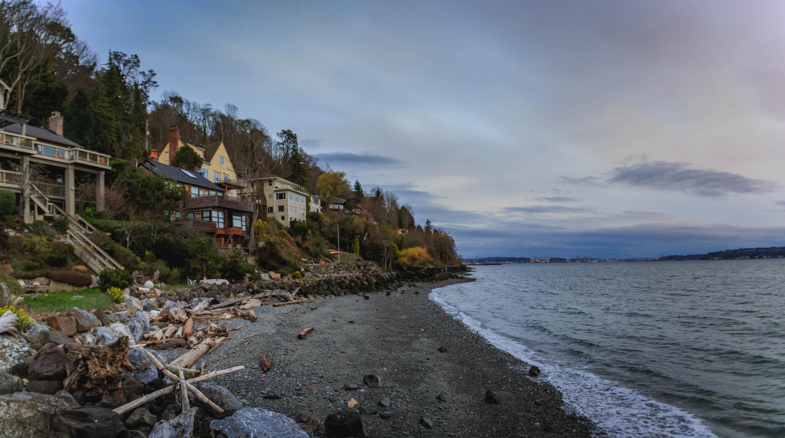 Houses sit atop a wooded hillside overlooking a rocky beach with driftwood along the shoreline, beside a calm body of water under a cloudy sky.