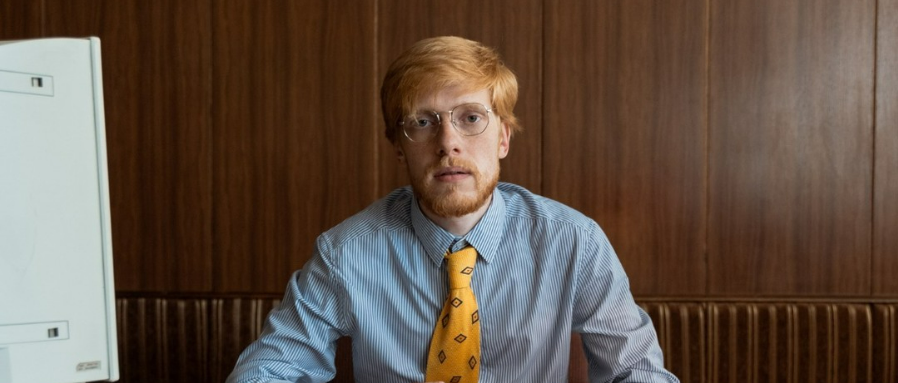A man with light skin, red hair, and glasses sits at a desk wearing a blue striped shirt and a yellow patterned tie, with a wooden wall and a computer monitor in the background.