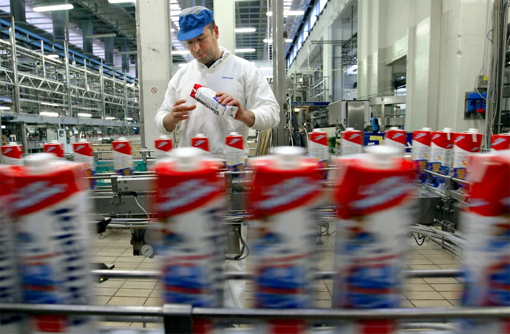 A worker in a white uniform and blue cap inspects a carton on a fast-moving production line in a brightly lit factory filled with equipment and rows of cartons.