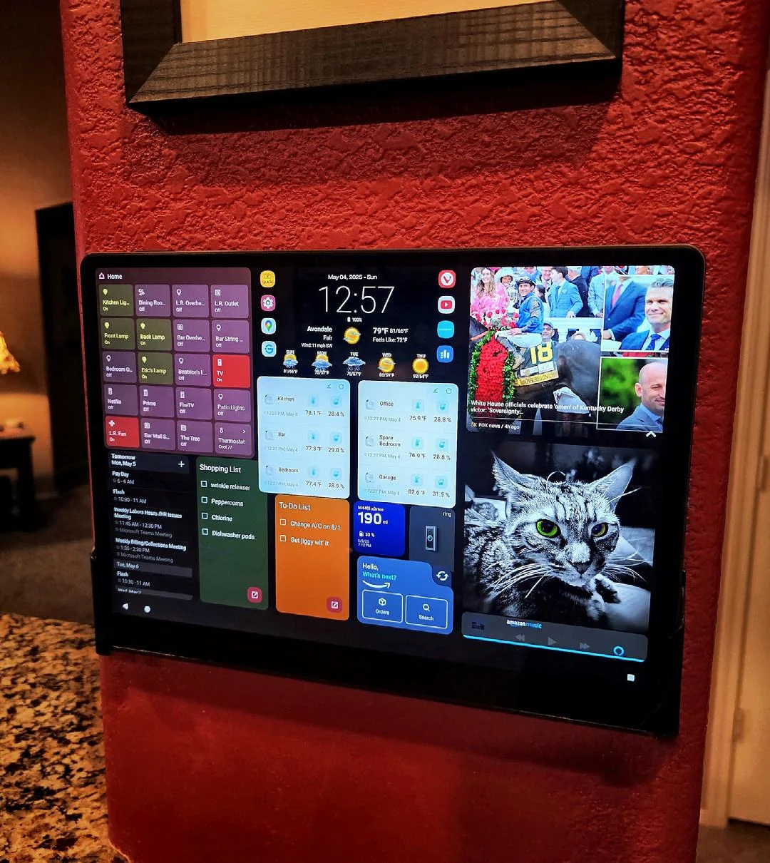 A tablet is mounted on a red wall, displaying a smart home dashboard with widgets for weather, calendar, news, home controls, and a photo of a gray cat in the bottom right corner.