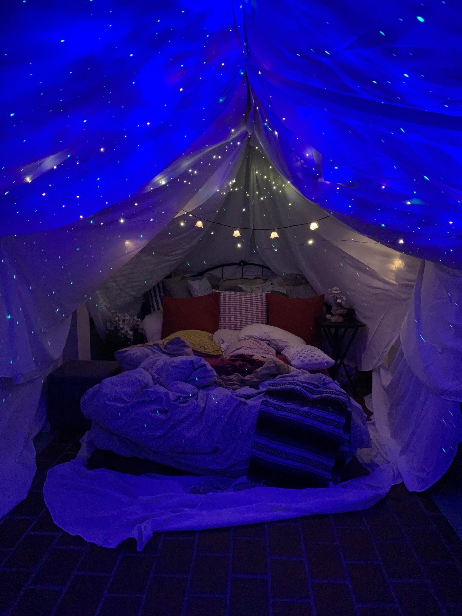 A cozy indoor fort made of white sheets is decorated with string lights and star projections on the ceiling. The bed inside is piled with pillows and blankets, creating a warm and inviting atmosphere.