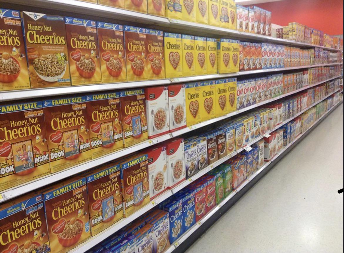 A grocery store aisle stocked with numerous colorful cereal boxes, including Cheerios and Honey Nut Cheerios, displayed neatly on long shelves stretching into the distance.