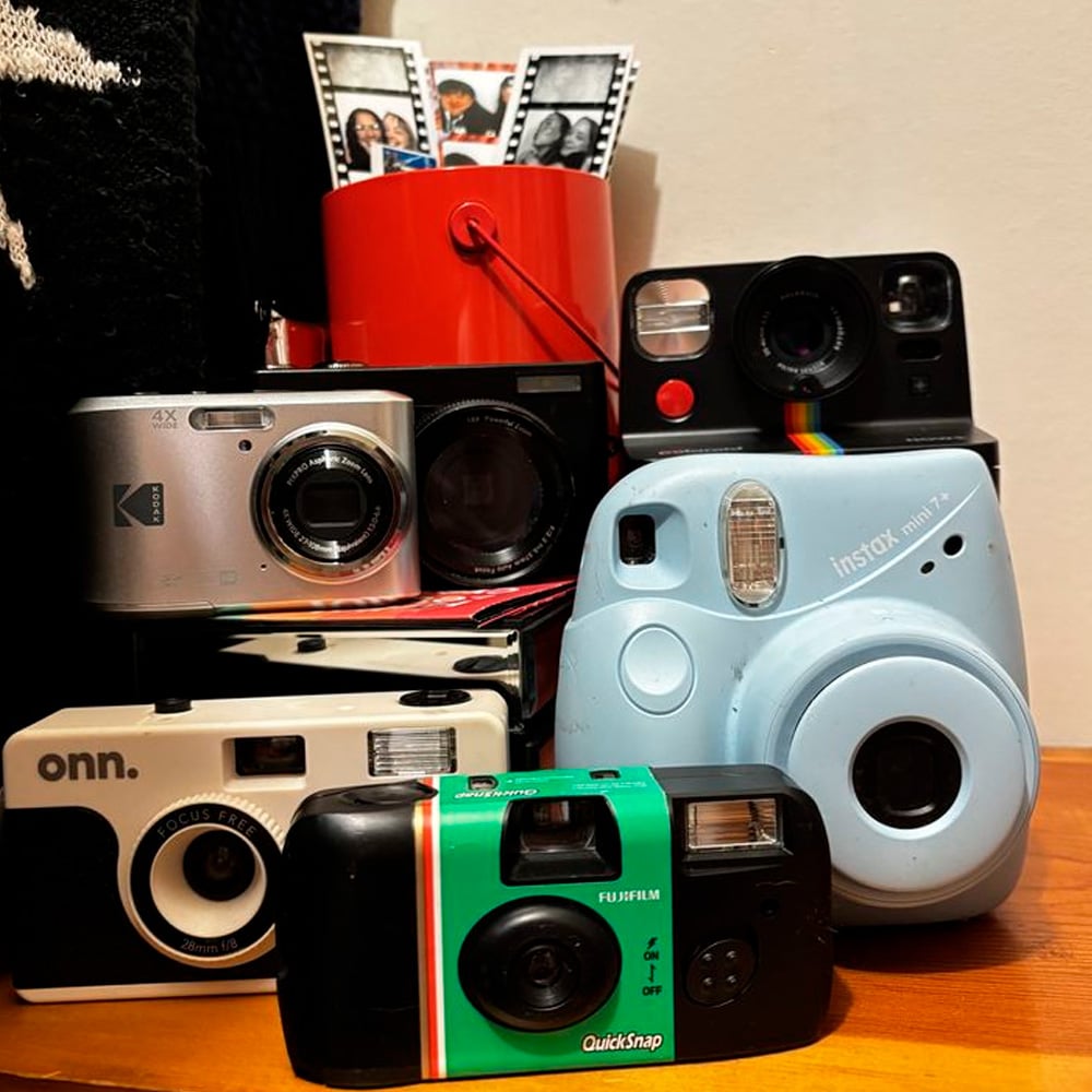 A collection of vintage and modern cameras stacked together, including film and instant models, with a red container holding black-and-white photo strips in the background.