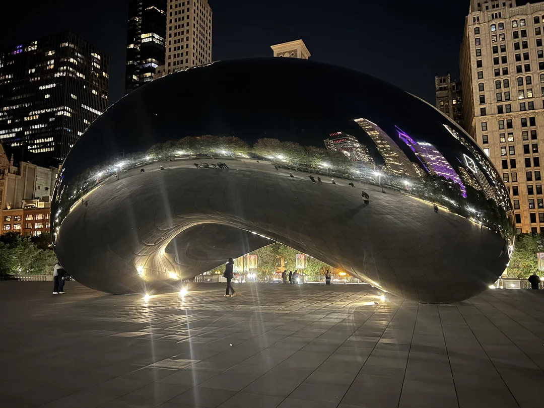 A nighttime view of Chicago’s Cloud Gate sculpture, also known as "The Bean," reflecting city lights, skyscrapers, and surrounding trees in its shiny surface. A few people are visible near the base.