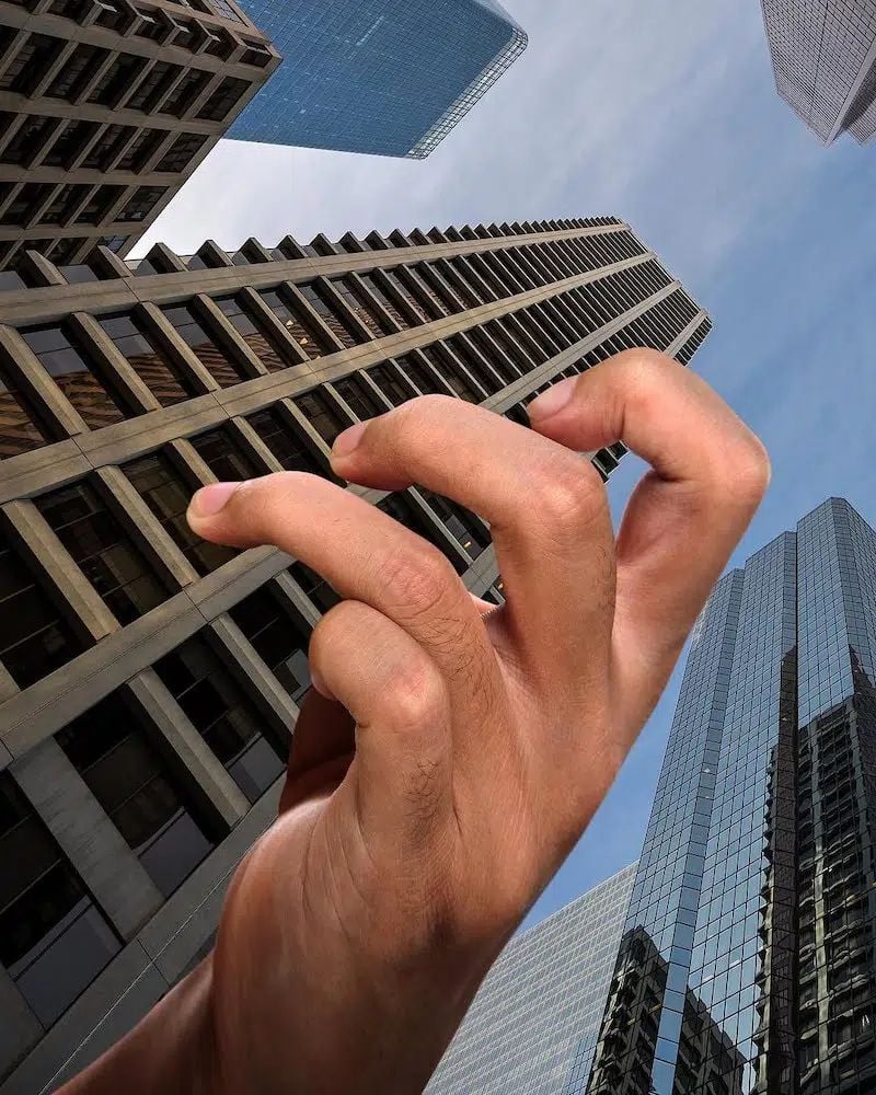 A large human hand appears oversized and out of proportion against a backdrop of tall, modern skyscrapers, giving the illusion of the hand towering above the buildings.