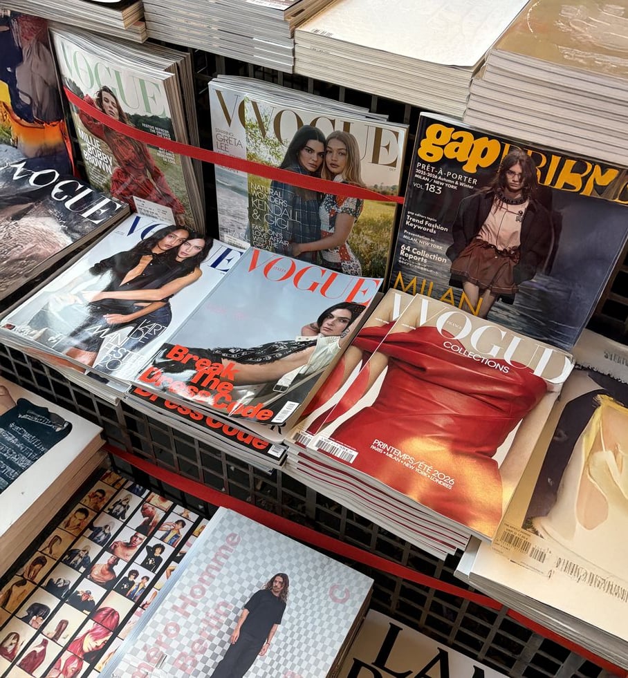 A magazine stand displays multiple issues of Vogue with fashion models on the covers, a Gap magazine, and other fashion publications arranged in neat, overlapping stacks.