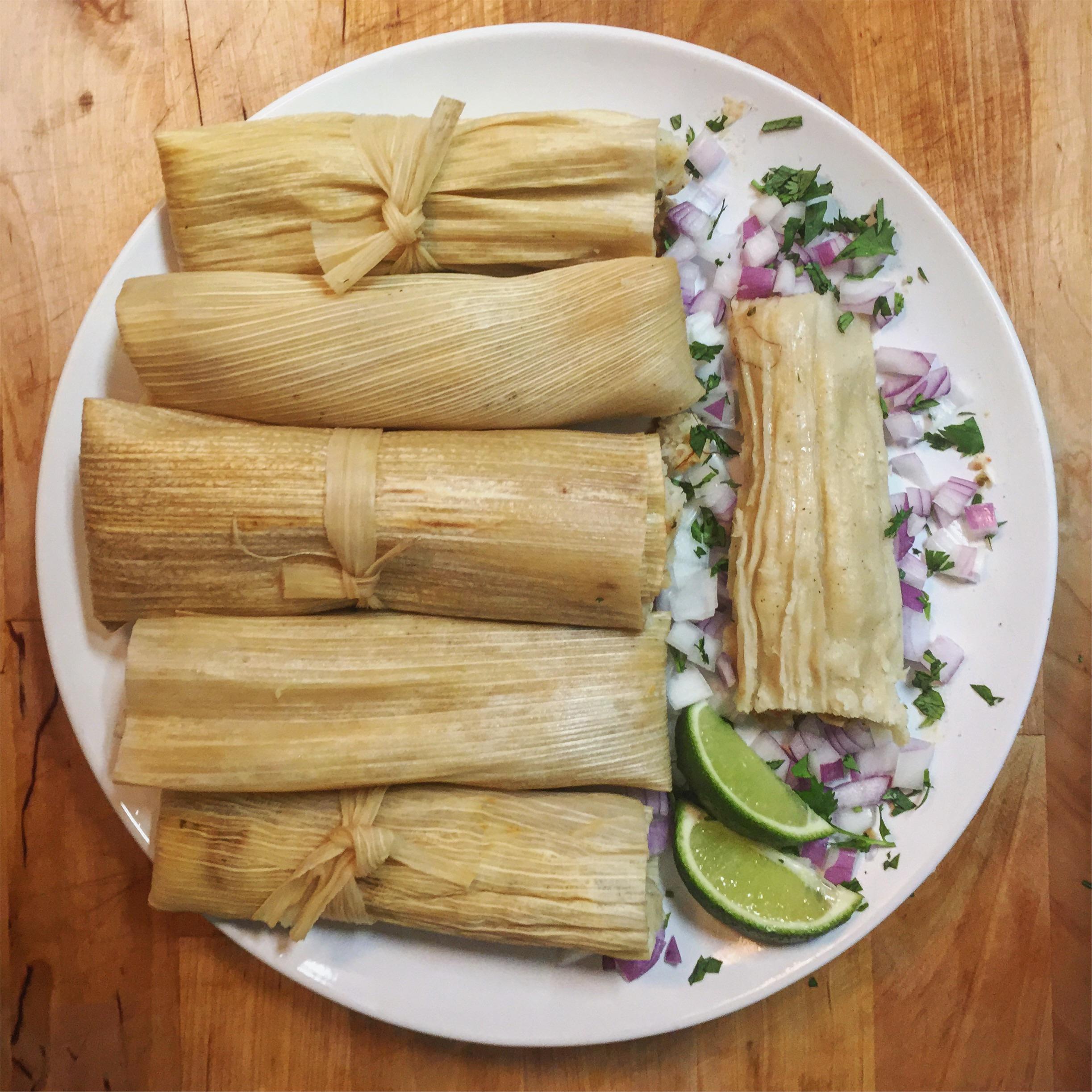 A plate with six tamales in corn husks, garnished with chopped onions, cilantro, and lime wedges on a wooden table. One tamale is unwrapped, showing its filling.