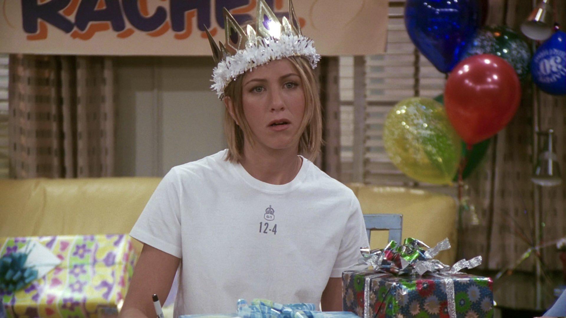 A woman wearing a crown sits at a table with wrapped presents and colorful balloons behind her. She looks surprised or confused. A banner with the name "Rachel" is partially visible in the background.