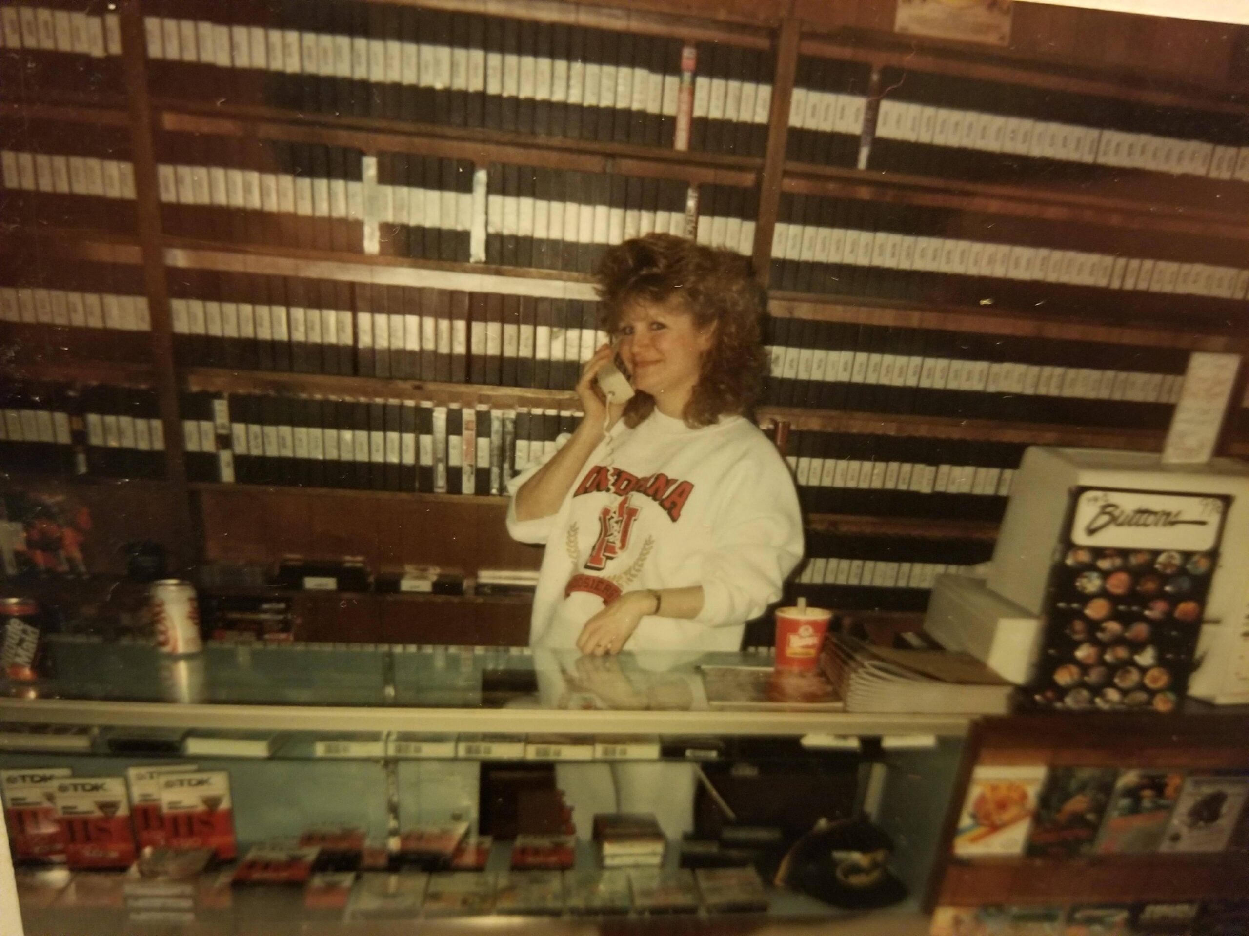 A woman with curly hair, wearing an Arizona sweatshirt, stands behind a store counter talking on the phone. Shelves behind her are filled with videotapes. A cash register and various items are on the counter.