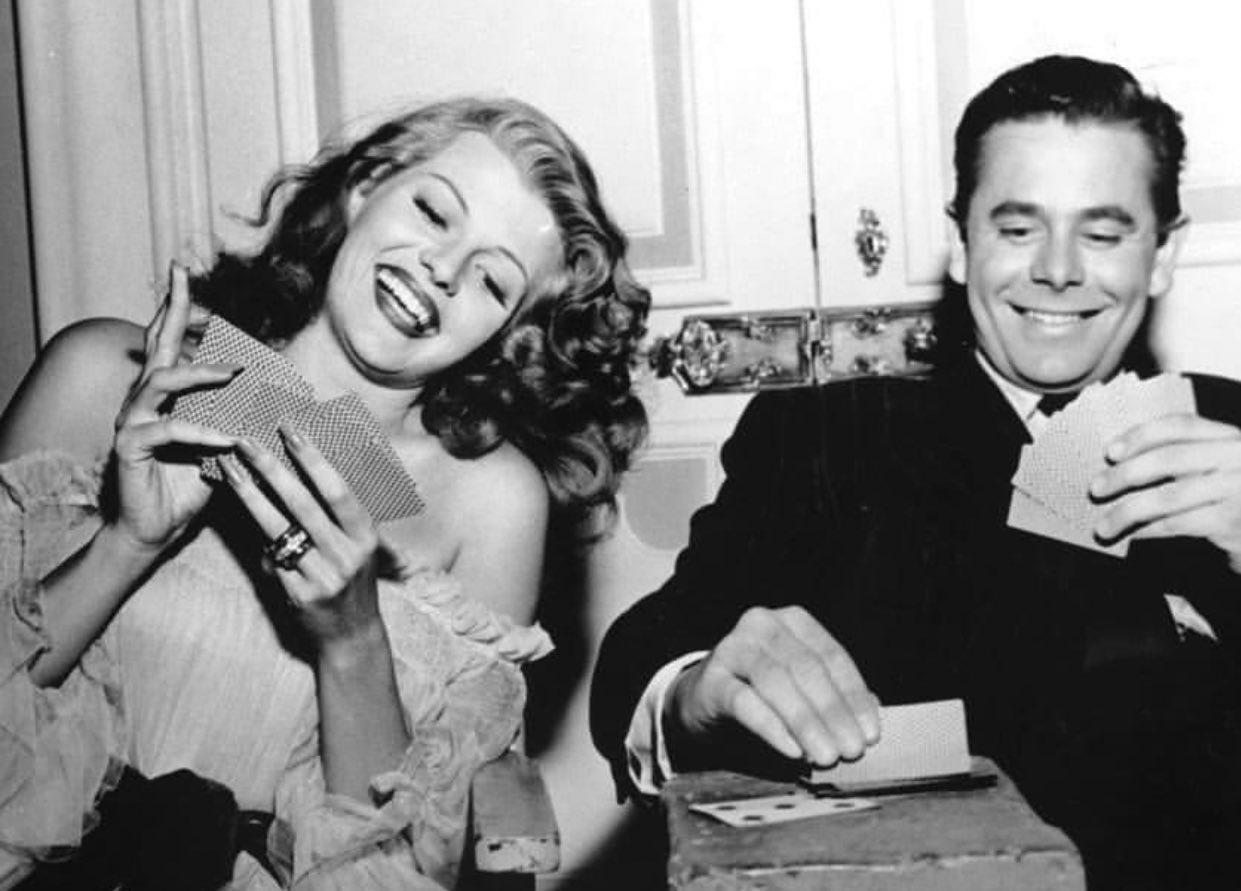 A woman and a man dressed formally are smiling and playing cards together. The woman holds her cards and laughs, while the man is also smiling, seated beside her at a small table. The setting looks elegant and vintage.