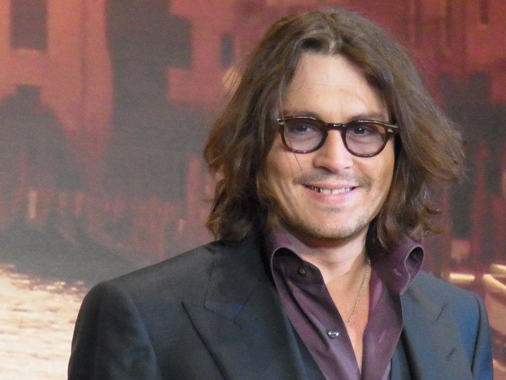 A man with shoulder-length brown hair and glasses, wearing a dark suit jacket over a maroon shirt, smiles while standing in front of a brownish blurred background.