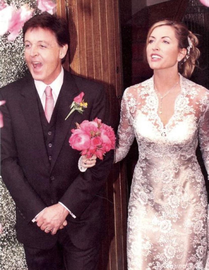 A smiling couple dressed in wedding attire stands at a doorway. The man wears a black suit with a pink tie and holds pink flowers; the woman wears a lacy white wedding gown and holds his arm. Pink flowers decorate the background.
