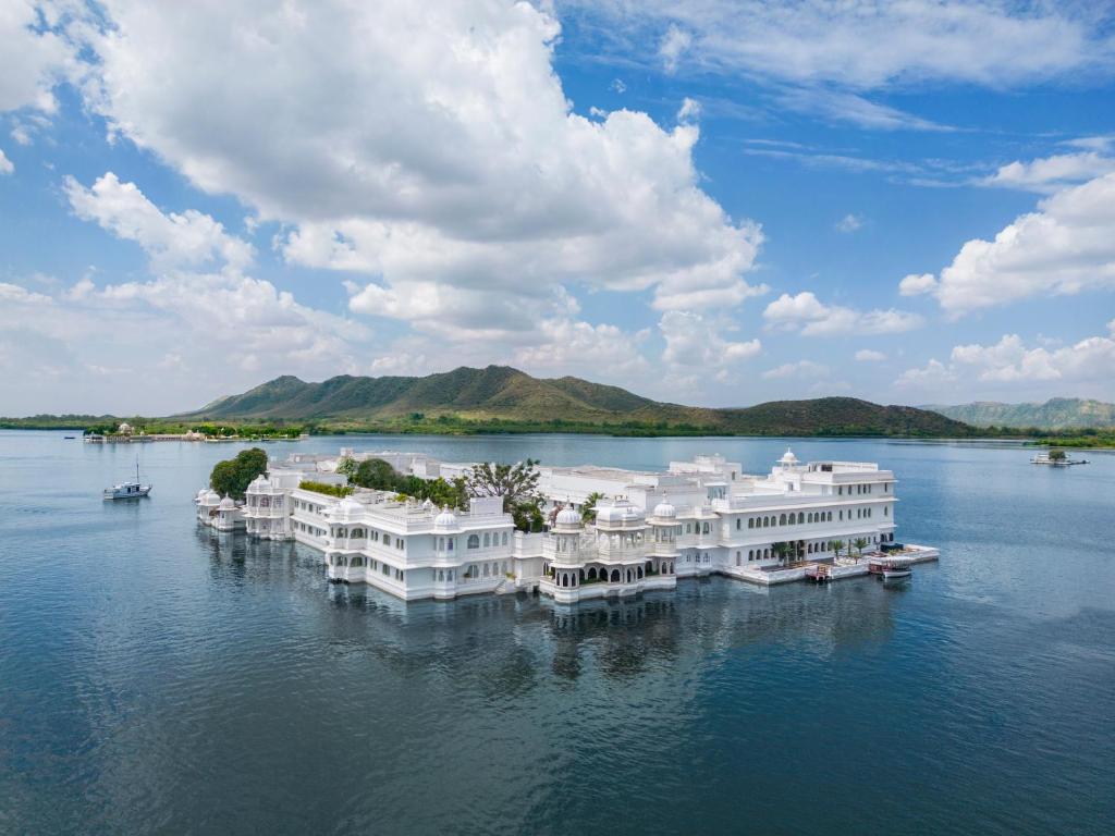 A white palace stands on a serene lake surrounded by mountains, under a blue sky with scattered clouds. Small boats are visible on the water near the palace.