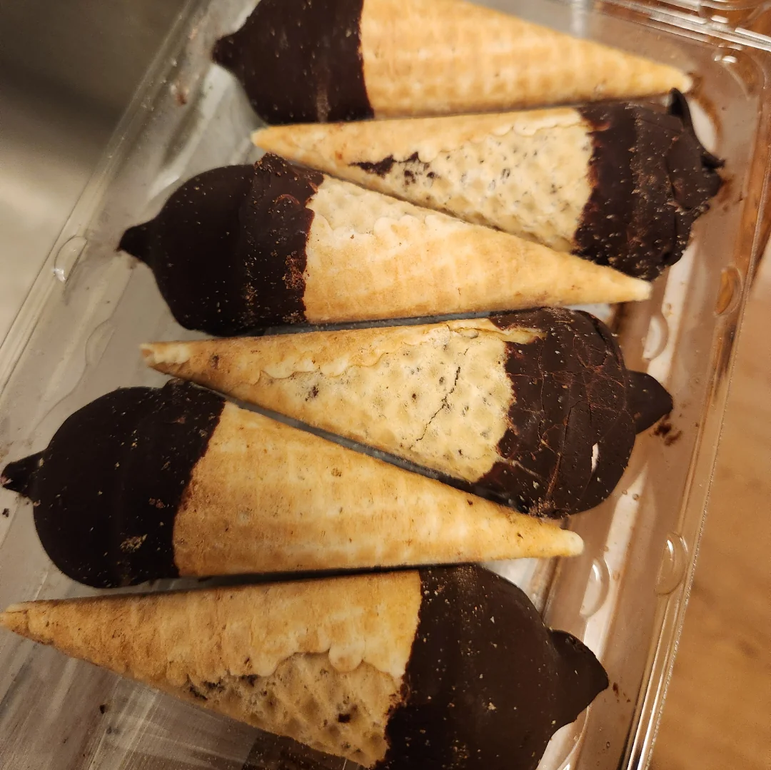 Six mini ice cream cones in a plastic container, each cone partially dipped in dark chocolate. The cones are arranged neatly in two rows, with the chocolate ends aligned in the same direction.