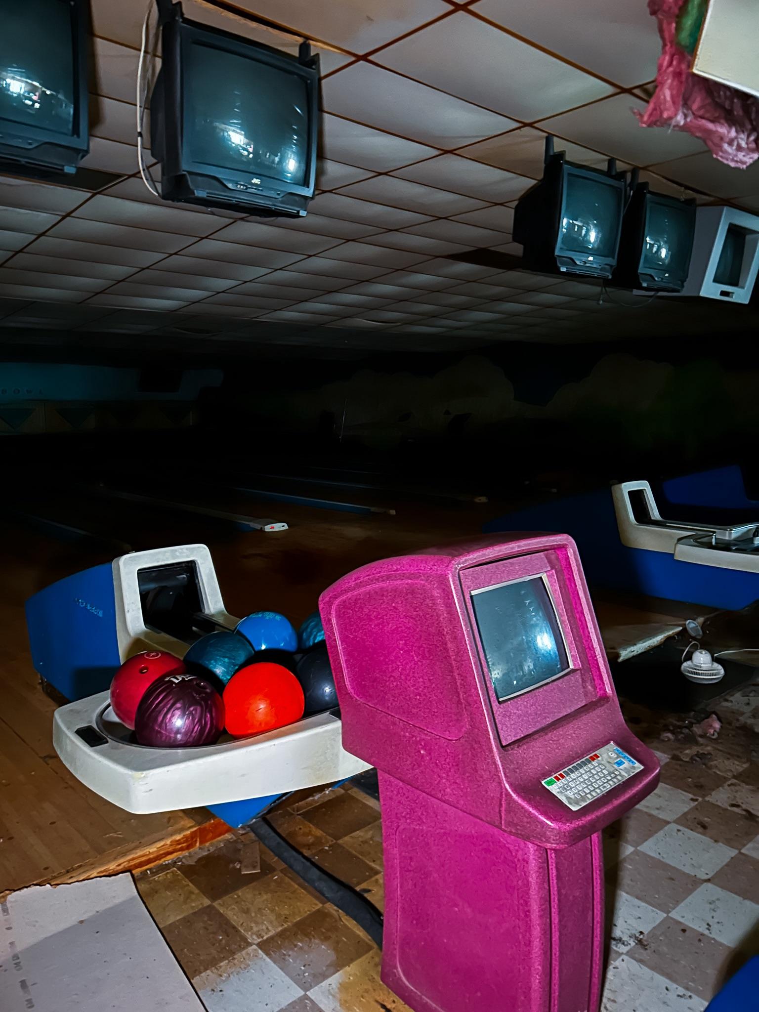 A dimly lit, possibly abandoned bowling alley with a pink scoring kiosk, multiple colorful bowling balls in a rack, and several old CRT monitors hanging from the ceiling. The area appears dusty and unused.